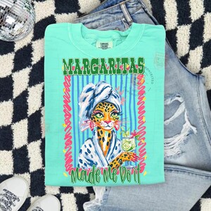 Pu&ograve; includere: T-shirt verde acqua con le parole "MARGARITAS" e "Made Me Do It" in verde e bianco. La maglietta presenta un'illustrazione di leopardo con asciugamano e accappatoio, che tiene in mano un cocktail. Il design &egrave; incorniciato da strisce rosa e blu.