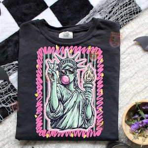 Puede incluir: Camiseta negra con un gr&aacute;fico de la Estatua de la Libertad con gafas de sol y soplando una burbuja de chicle. La Estatua de la Libertad sostiene una antorcha y hace el signo de la paz. El dise&ntilde;o est&aacute; delineado en rosa y verde.