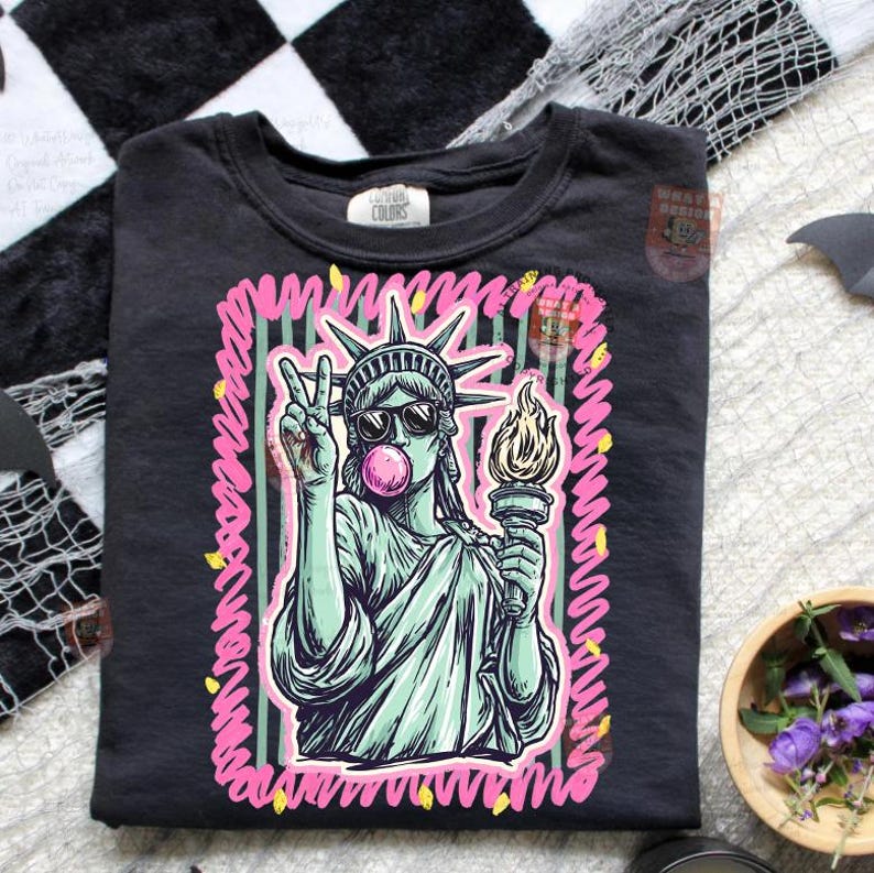Puede incluir: Camiseta negra con un gr&aacute;fico de la Estatua de la Libertad con gafas de sol y soplando una burbuja de chicle. La Estatua de la Libertad sostiene una antorcha y hace el signo de la paz. El dise&ntilde;o est&aacute; delineado en rosa y verde.