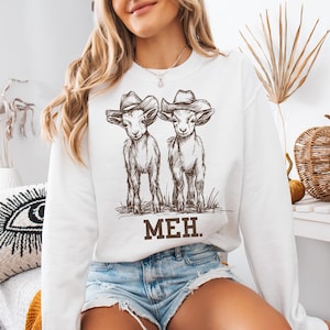 May include: White sweatshirt with a graphic of two goats wearing cowboy hats. The text "MEH." is printed below the goats.