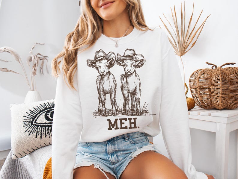 May include: White sweatshirt with a graphic of two goats wearing cowboy hats. The text "MEH." is printed below the goats.
