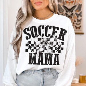Puede incluir: Sudadera blanca de manga larga con texto negro que dice "Soccer Mama" y un patr&oacute;n de cuadros blanco y negro con un lazo de bal&oacute;n de f&uacute;tbol.