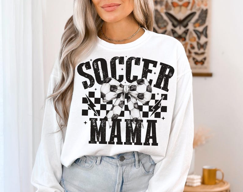 Puede incluir: Sudadera blanca de manga larga con texto negro que dice "Soccer Mama" y un patr&oacute;n de cuadros blanco y negro con un lazo de bal&oacute;n de f&uacute;tbol.