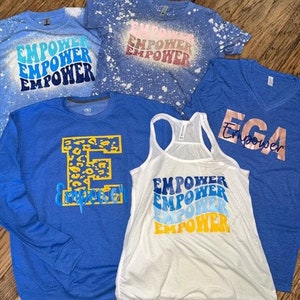 May include: Five blue and white shirts with the word "Empower" printed on them in different styles. One shirt has a leopard print "E" and the words "Empower" underneath. One shirt has a white background with blue and yellow "Empower" text. One shirt has a blue background with white "Empower" text. One shirt has a blue background with white "Empower" text and a white splatter pattern. One shirt has a blue background with gold "Empower" text.