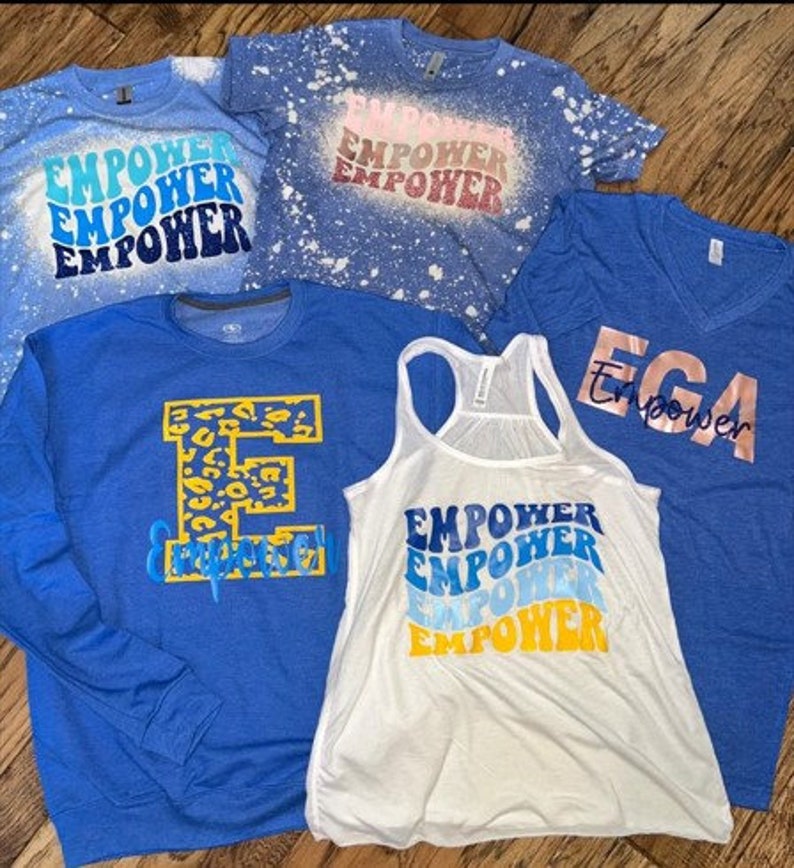May include: Five blue and white shirts with the word "Empower" printed on them in different styles. One shirt has a leopard print "E" and the words "Empower" underneath. One shirt has a white background with blue and yellow "Empower" text. One shirt has a blue background with white "Empower" text. One shirt has a blue background with white "Empower" text and a white splatter pattern. One shirt has a blue background with gold "Empower" text.