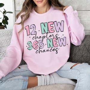 K&ouml;nnte beinhalten: Rosafarbener Pullover mit dem Text "12 Neue Kapitel 365 Neue Chancen" in bunten, umrandeten Buchstaben.