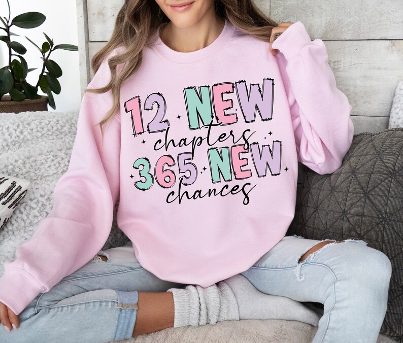 K&ouml;nnte beinhalten: Rosafarbener Pullover mit dem Text "12 Neue Kapitel 365 Neue Chancen" in bunten, umrandeten Buchstaben.