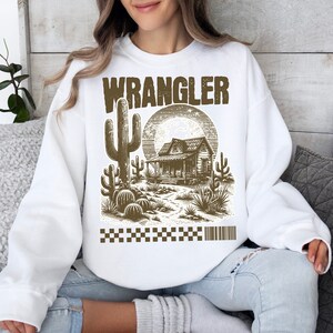 Pode incluir: Camisola branca com uma estampa gr&aacute;fica marrom de uma cena de deserto com um cacto, uma cabana e um p&ocirc;r do sol. O texto "WRANGLER" &eacute; impresso em marrom acima do gr&aacute;fico.