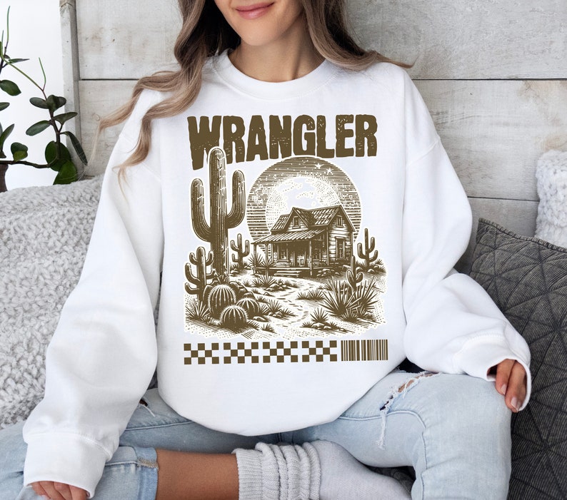 Pode incluir: Camisola branca com uma estampa gr&aacute;fica marrom de uma cena de deserto com um cacto, uma cabana e um p&ocirc;r do sol. O texto "WRANGLER" &eacute; impresso em marrom acima do gr&aacute;fico.