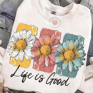 May include: White sweatshirt featuring three colorful daisy flowers in yellow, coral, and teal. The flowers are set against rectangular backgrounds. The phrase "Life is Good" is written in black script below the flowers.