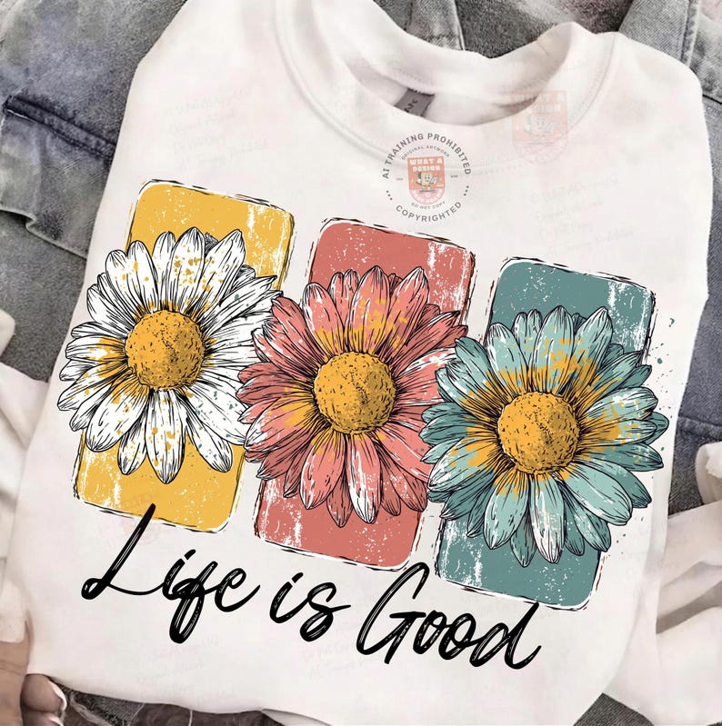 May include: White sweatshirt featuring three colorful daisy flowers in yellow, coral, and teal. The flowers are set against rectangular backgrounds. The phrase "Life is Good" is written in black script below the flowers.