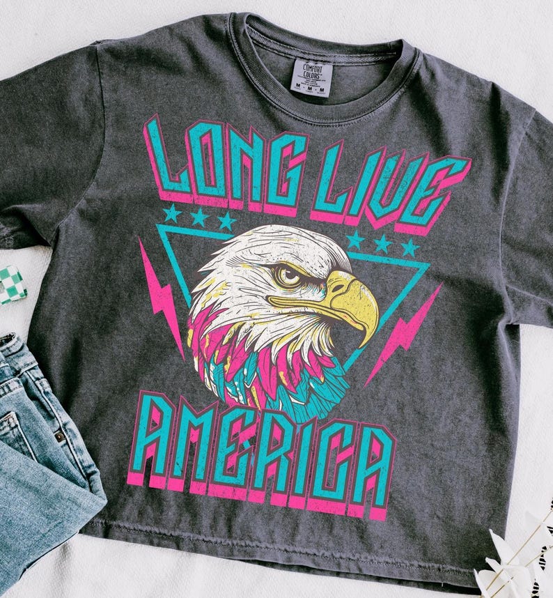 May include: A dark grey t-shirt with a vintage-style graphic of an eagle with the words "Long Live America" in pink and blue lettering.