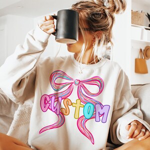 K&ouml;nnte beinhalten: Cremefarbenes Sweatshirt mit einer gro&szlig;en, bunten Schleifen-Grafik und dem Wort "CUSTOM" in Buchstaben. Die Schleife hat rosa, blaue und gelbe Streifen. Das Sweatshirt ist ein l&auml;ssiger, bequemer Stil.