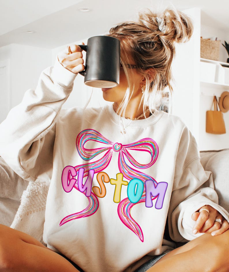 K&ouml;nnte beinhalten: Cremefarbenes Sweatshirt mit einer gro&szlig;en, bunten Schleifen-Grafik und dem Wort "CUSTOM" in Buchstaben. Die Schleife hat rosa, blaue und gelbe Streifen. Das Sweatshirt ist ein l&auml;ssiger, bequemer Stil.