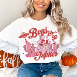 K&ouml;nnte beinhalten: Wei&szlig;er Pullover mit einem pinkfarbenen und roten Grafikdesign, das einen Cowboy zeigt, der ein UFO reitet, mit dem Text "Boys Go To Jupiter".