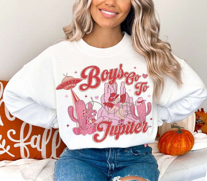 K&ouml;nnte beinhalten: Wei&szlig;er Pullover mit einem pinkfarbenen und roten Grafikdesign, das einen Cowboy zeigt, der ein UFO reitet, mit dem Text "Boys Go To Jupiter".
