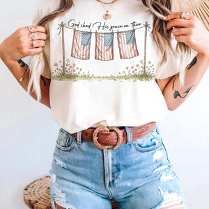 May include: Cream-colored t-shirt featuring a graphic of three American flags hanging on a clothesline, with the text "God shed His grace on thee." The flags are red, white, and blue. The shirt is paired with blue denim shorts and a brown belt.