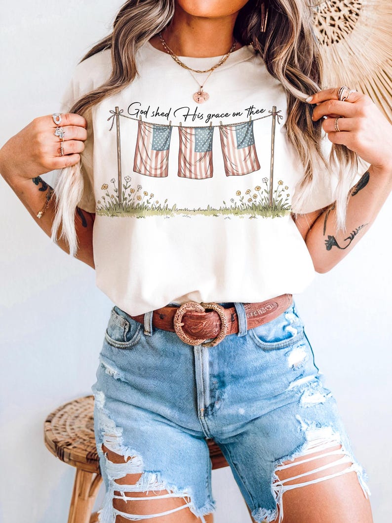 May include: Cream-colored t-shirt featuring a graphic of three American flags hanging on a clothesline, with the text "God shed His grace on thee." The flags are red, white, and blue. The shirt is paired with blue denim shorts and a brown belt.