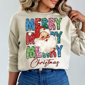 Op de afbeelding: Een beige sweatshirt met een feestelijk kerstontwerp. Het ontwerp bevat de woorden "Merry" drie keer herhaald in kleurrijke glitterletters, met een vintage illustratie van de kerstman in het midden. De woorden "Christmas" zijn in rode glitter geschreven onder de kerstman-illustratie.