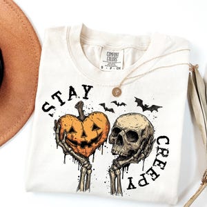 Puede incluir: Camiseta color crema con un gr&aacute;fico tem&aacute;tico de Halloween. El dise&ntilde;o presenta una calabaza sujetada por manos esquel&eacute;ticas, una calavera y murci&eacute;lagos. El texto "STAY CREEPY" est&aacute; impreso alrededor del dise&ntilde;o. Un collar dorado est&aacute; sobre la camiseta.