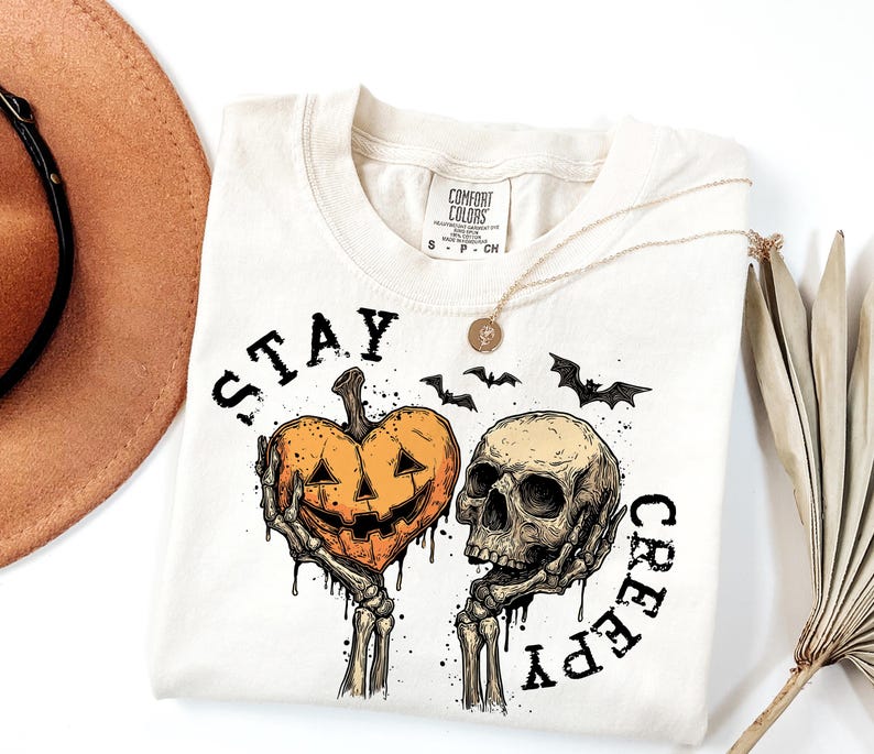 Puede incluir: Camiseta color crema con un gr&aacute;fico tem&aacute;tico de Halloween. El dise&ntilde;o presenta una calabaza sujetada por manos esquel&eacute;ticas, una calavera y murci&eacute;lagos. El texto "STAY CREEPY" est&aacute; impreso alrededor del dise&ntilde;o. Un collar dorado est&aacute; sobre la camiseta.