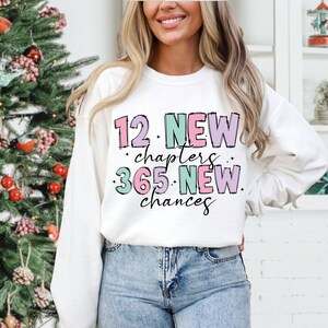 K&ouml;nnte beinhalten: Wei&szlig;er Pullover mit einem pinkfarbenen, violetten und gr&uuml;nen Grafikdesign, auf dem "12 neue Kapitel 365 neue Chancen" steht.