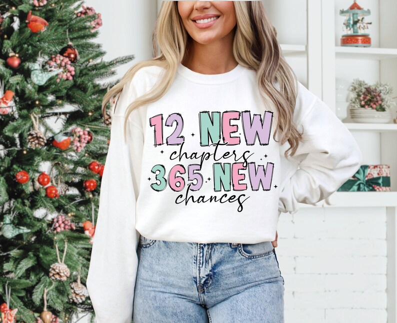 K&ouml;nnte beinhalten: Wei&szlig;er Pullover mit einem pinkfarbenen, violetten und gr&uuml;nen Grafikdesign, auf dem "12 neue Kapitel 365 neue Chancen" steht.