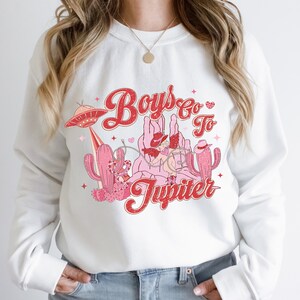 K&ouml;nnte beinhalten: Wei&szlig;er Pullover mit einem roten und rosafarbenen Grafikdesign mit dem Text "Boys Go To Jupiter" mit einem Cowboyhut, einem Kaktus und einem UFO.