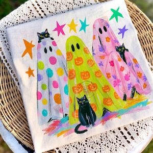 May include: A white t-shirt featuring a graphic of three ghosts draped in patterned sheets, each accompanied by a black cat. The ghosts are decorated with colorful polka dots, orange pumpkins, and pink floral prints, surrounded by bright stars.