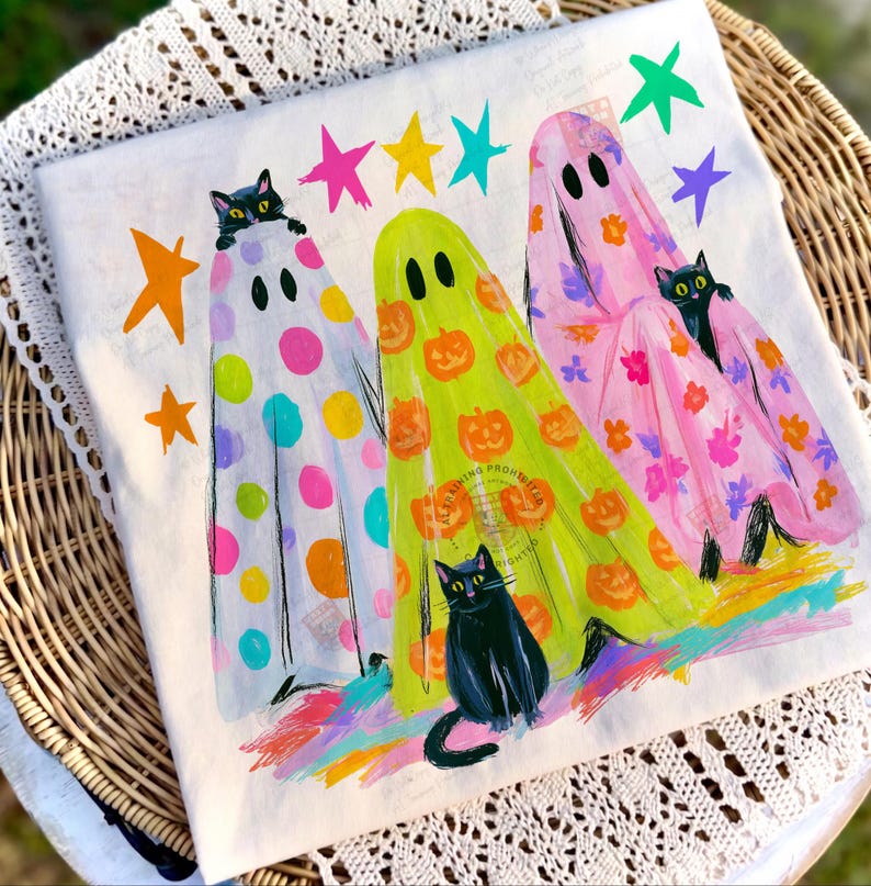 May include: A white t-shirt featuring a graphic of three ghosts draped in patterned sheets, each accompanied by a black cat. The ghosts are decorated with colorful polka dots, orange pumpkins, and pink floral prints, surrounded by bright stars.