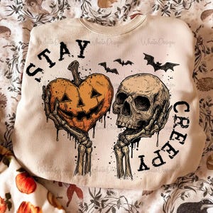 Puede incluir: Sudadera crema con dise&ntilde;o de Halloween. El gr&aacute;fico presenta una calabaza en forma de coraz&oacute;n y una calavera, sostenidas por manos esquel&eacute;ticas, con las palabras "STAY CREEPY" y murci&eacute;lagos. Un dise&ntilde;o espeluznante y festivo.