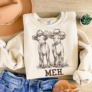 May include: A cream-colored sweatshirt with a graphic of two goats wearing cowboy hats. The text "MEH." is printed below the goats.