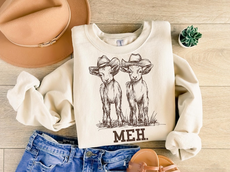 May include: A cream-colored sweatshirt with a graphic of two goats wearing cowboy hats. The text "MEH." is printed below the goats.