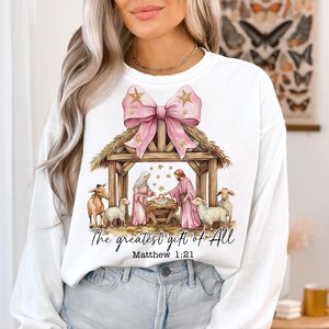 May include: White long-sleeve sweatshirt with a pink bow and a nativity scene graphic. The graphic includes a manger, a baby Jesus, Mary, Joseph, and animals. The text "The greatest gift of all" and "Matthew 1:21" is printed below the graphic.