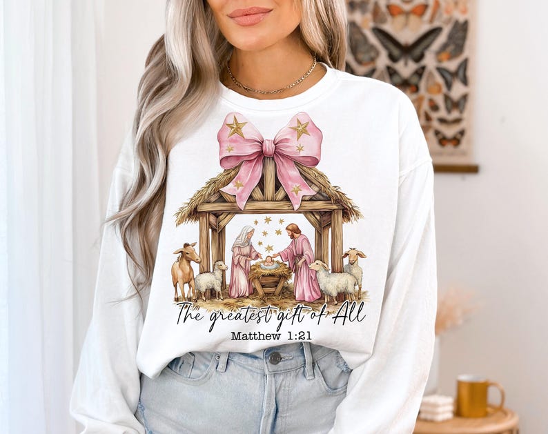 May include: White long-sleeve sweatshirt with a pink bow and a nativity scene graphic. The graphic includes a manger, a baby Jesus, Mary, Joseph, and animals. The text "The greatest gift of all" and "Matthew 1:21" is printed below the graphic.