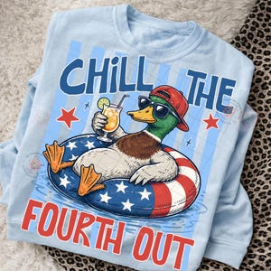 May include: Light blue sweatshirt with a graphic of a duck wearing sunglasses and a red cap, relaxing in a patriotic float with a cocktail. Text reads "CHILL THE FOURTH OUT" in blue and red.