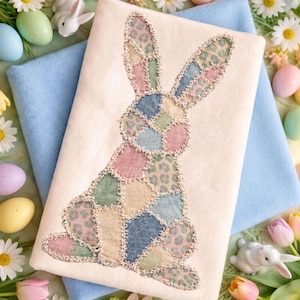 Puede incluir: Un cojín crema con un diseño de conejito de patchwork en colores pastel. El conejito está hecho de varios cuadrados de tela en rosa, azul, verde y beige, con un estampado de leopardo. El cojín está rodeado de huevos de Pascua, flores y figuritas de conejos.