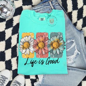 May include: A turquoise t-shirt with a floral design featuring three daisies in yellow, pink, and blue. The text "Life is Good" is printed below the flowers. The shirt is folded on a black and white checkered surface.