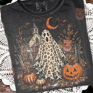 May include: A charcoal gray t-shirt featuring a graphic print of a ghost with a leopard print pattern, a church, a crescent moon, and pumpkins. The shirt has a crew neckline and a Comfort Colors brand label printed on the inside of the collar.