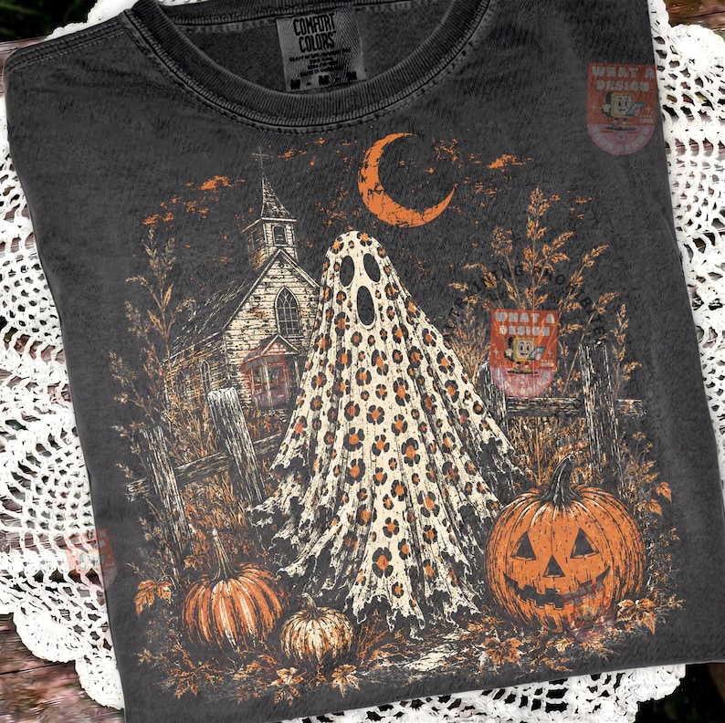 May include: A charcoal gray t-shirt featuring a graphic print of a ghost with a leopard print pattern, a church, a crescent moon, and pumpkins. The shirt has a crew neckline and a Comfort Colors brand label printed on the inside of the collar.