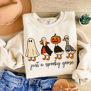 May include: Cream-colored sweatshirt with a Halloween-themed graphic. The design features four cartoon geese dressed as a ghost, vampire, jack-o'-lantern, and skeleton. The text "just a spooky goose" is written below the geese. The sweatshirt is paired with blue jeans and a tan hat.