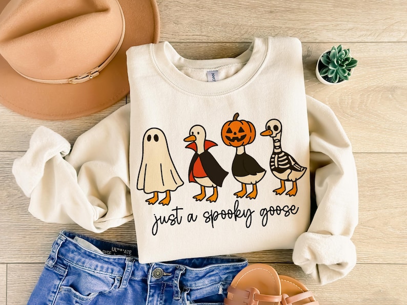 May include: Cream-colored sweatshirt with a Halloween-themed graphic. The design features four cartoon geese dressed as a ghost, vampire, jack-o'-lantern, and skeleton. The text "just a spooky goose" is written below the geese. The sweatshirt is paired with blue jeans and a tan hat.
