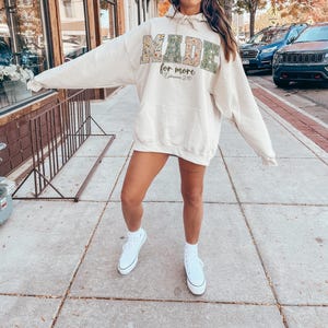 May include: Cream-colored hoodie with the words "MADE for more" in a floral and green design. The hoodie has a front pocket and a drawstring hood. The text "Ephesians 2:10" is below the main text. The person is wearing white sneakers.