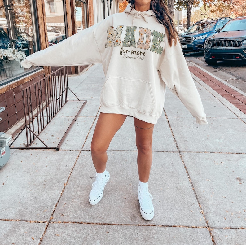 May include: Cream-colored hoodie with the words "MADE for more" in a floral and green design. The hoodie has a front pocket and a drawstring hood. The text "Ephesians 2:10" is below the main text. The person is wearing white sneakers.