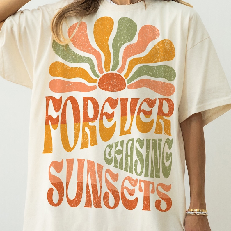 May include: Cream-colored t-shirt featuring a retro-style graphic. The design includes a stylized sunburst in orange, yellow, and green, with the words "FOREVER CHASING SUNSETS" in a matching vintage font. The shirt has short sleeves.