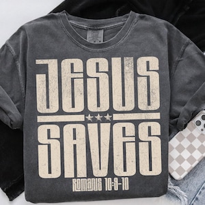 Puede incluir: Una camiseta gris oscura con un gráfico blanco desgastado que dice "Jesus Saves" con tres estrellas sobre la palabra "Saves" y el texto "Romanos 10:9-10" debajo.