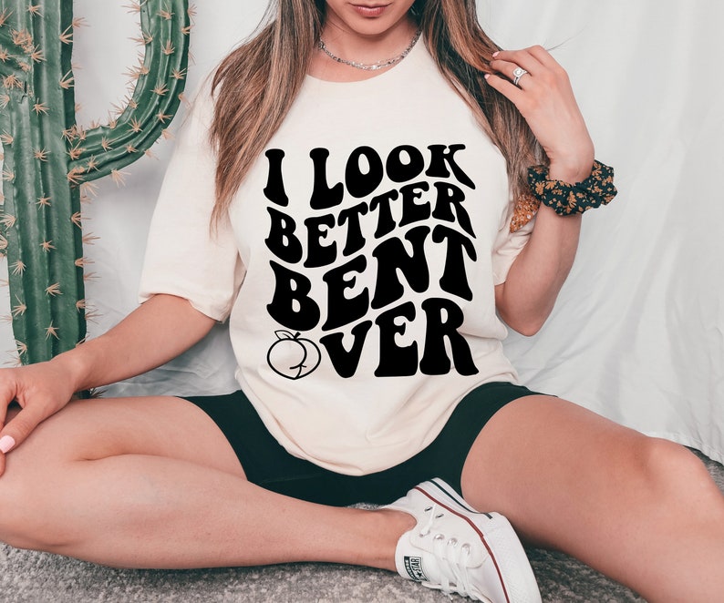 May include: A white t-shirt with black text that reads "I look better bent over" in a retro font. A peach graphic is below the text.