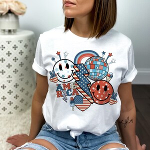 May include: White t-shirt with a red, white and blue graphic design featuring smiley faces, stars, a lightning bolt and the word "AMERICA".