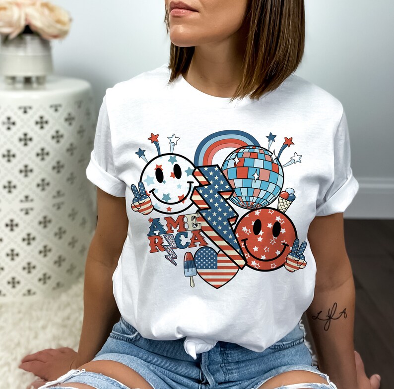 May include: White t-shirt with a red, white and blue graphic design featuring smiley faces, stars, a lightning bolt and the word "AMERICA".