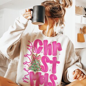 May include: Cream-colored sweatshirt with the words "Merry Christmas" in pink, featuring a Christmas tree graphic. The tree is green with a pink bow and gold accents. The sweatshirt is a casual, festive garment.